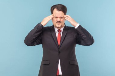 Bıyıklı hasta adam kendini iyi hissetmiyor, baş ağrısı çekiyor, kaşlarını çatıyor, şakaklara masaj yapıyor, kırmızı kravatlı siyah takım elbise giyiyor. Açık mavi arkaplanda izole edilmiş kapalı stüdyo çekimi.