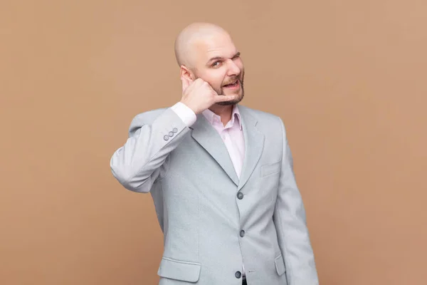 You contact us. Portrait of smiling bald bearded man making gesture with fingers dial my number or call me back, wearing gray jacket. Indoor studio shot isolated on brown background.