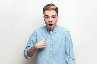 Şok olmuş, şaşkın, kot gömlek giyen, kendini gösteren, kameraya büyük gözlerle bakan ve ağzı açık bir adamın portresi. Gri arka planda izole edilmiş kapalı stüdyo görüntüleri.