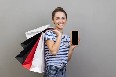 Alışveriş çantaları ve cep telefonu taşıyan mutlu bir kadının portresi, online sipariş için reklam başvurusu, mal satın alma ve teslimatı. Gri arka planda izole edilmiş kapalı stüdyo görüntüleri.