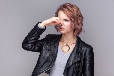 Kısa saç stili olan üzgün bir kadının portresi korkunç bir koku, burnu sıkıyor, yüzünü buruşturuyor, siyah deri ceket giyiyor. Kapalı alanda çekilen çekim gri arka planda izole edildi.