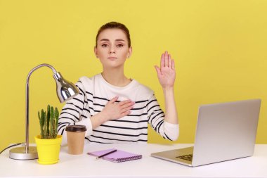 Sorumluluk sahibi, dürüst bir kadın işyerinde oturuyor ve söz vermek için elini kaldırıyor. Güvenilir, sadık bir ifadeyle yemin ediyor. Sarı arka planda kapalı stüdyo çekimleri.