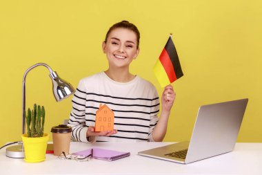 Alman bayrağı taşıyan, elinde kağıt evle iş yerinde oturan, konaklama satın almaktan memnun bir kadın. Sarı arka planda kapalı stüdyo çekimleri.