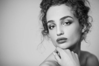 Çekici, güzel bir kadın mankenin siyah beyaz portresi. Siyah kıvırcık saçlı ve makyajlı, kameraya bakıyor. Gri arka planda izole edilmiş kapalı stüdyo görüntüleri.