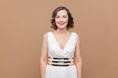 Gülümseyen, mutlu, iyimser, dalgalı saçlı, beyaz elbiseli, kameraya bakan orta yaşlı kadının portresi. Açık kahverengi arka planda kapalı stüdyo çekimleri.