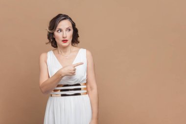 Şaşırmış orta yaşlı kadın portresi dalgalı saçlarıyla reklam için fotokopi alanını işaret ediyor, beyaz elbise giyiyor. Açık kahverengi arka planda kapalı stüdyo çekimleri.