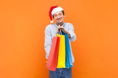 Renkli alışveriş torbaları tutan, mutlu bir gülümsemeyle kameraya bakan, açık mavi gömlek giyen ve Noel Baba şapkası takan mutlu bir adamın portresi. Turuncu arka planda kapalı stüdyo çekimleri.