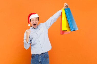 Son derece mutlu genç bir adamın portresi sıkılmış yumruğu ve alışveriş torbaları, başarılı alışverişi kutlayan açık mavi gömlek ve Noel Baba şapkası giyen. Turuncu arka planda kapalı stüdyo çekimleri.