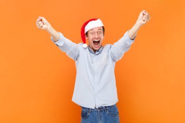 Son derece mutlu bir adamın portresi kış tatilini kutlayan mutlu bir şekilde bağıran, kollarını kaldırmış, Noel Baba şapkası takan. Turuncu arka planda kapalı stüdyo çekimleri.