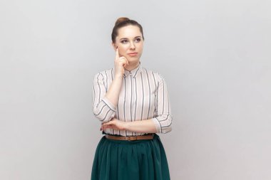 Çizgili gömlek ve yeşil etek giyen düşünceli bir kadının portresi. Çenesini tutuyor, başka tarafa bakıyor, geleceğini planlıyor, işini düşünüyor. Gri arka planda izole edilmiş kapalı stüdyo görüntüleri.