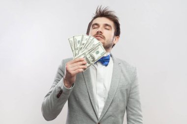 Zengin gururlu sakallı adamın portresi dolar banknotları kokluyor, yüksek maaştan memnun, gri takım elbise giyiyor ve mavi papyon takıyor. Gri arka planda izole edilmiş kapalı stüdyo görüntüleri.