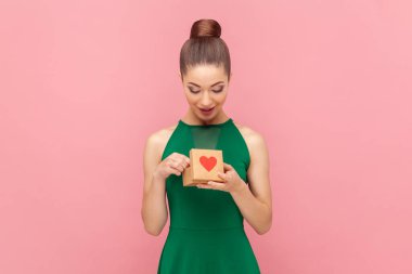 Pozitif iyimser iyimser kadının portresi küçük hediye kutusuyla duruyor, içine bakıyor, hediyesine hayran, yeşil elbise giyiyor. Pembe arka planda kapalı stüdyo çekimleri.
