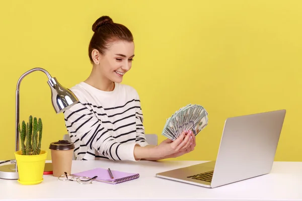 Satisfied rich woman office manager showing hundred dollar bills to laptop display, sitting at workplace, satisfied about big profit. Indoor studio studio shot isolated on yellow background.