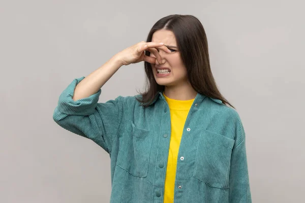 Woman pinching nose, stop breathing bad odor, disgusted by smell of farting, her grimace expressing repulsion, gross, wearing casual style jacket. Indoor studio shot isolated on gray background.