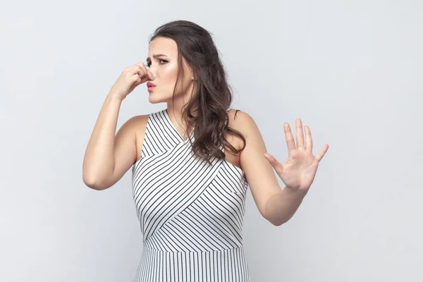 Displeased woman covers nose with hand, smells something awful, pinches nose, frowns in displeasure, sees pile of garbage, wearing striped dress. Indoor studio shot isolated on gray background.