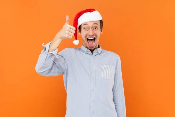 stock image Portrait of excited positive man standing showing thumb up, demonstrates like gesture, looking at camera, wearing light blue shirt and Santa Claus hat. Indoor studio shot isolated on orange background