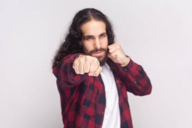 Uzun kıvırcık saçlı, kareli kırmızı gömlekli agresif sakallı adam yumruklarını dövüşmeye hazır tutuyor ve adaletsizliğe ve şiddete karşı kendini savunuyor. Gri arka planda izole edilmiş kapalı stüdyo görüntüleri.
