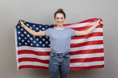 Çizgili tişört giyen, büyük Amerikan bayrağı tutan mutlu ve genç bir kadının portresi. Olumlu duyguları ve mutluluğu ifade ediyor. Gri arka planda izole edilmiş kapalı stüdyo görüntüleri.