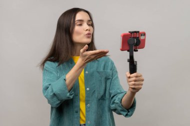 Akıllı telefonu elinde sabit kamerayla tutan romantik bir kadın, canlı yayın yapan ya da selfie çeken bir blog yazarı, hava öpücüğü yollayan, günlük tarz ceket giyen. Gri arka planda izole edilmiş kapalı stüdyo görüntüleri