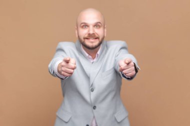 Seni işaret eden neşeli kel sakallı adamın portresi, neşeli bir ifadeye sahip, dişlek gülümsemeli kameraya bakıyor, gri ceket giyiyor. Kahverengi arka planda kapalı stüdyo çekimleri.