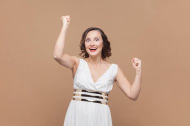 Son derece mutlu orta yaşlı kadın portresi, dalgalı saçları olan, yumruklarını sallayan ve mutlu bir şekilde bağıran, zaferini kutlayan, beyaz elbise giyen. Açık kahverengi arka planda kapalı stüdyo çekimleri.