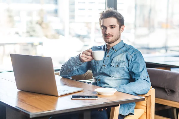 Dizüstü bilgisayarda çalışan, sunum izleyip kahve içen, kot pantolonlu, serbest çalışan sakin genç bir adamın portresi. Büyük pencerenin yanında, kafenin arka planında..