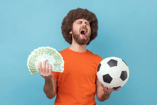 穿着橙色T恤 拿着足球和几百张欧元钞票 兴高采烈地尖叫着 投注着 还赢了的兴奋的黑人发型男人 室内拍摄被蓝色背景隔离 — 图库照片