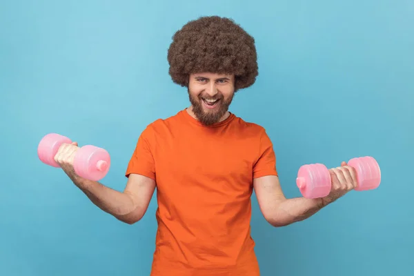 Portrait of strong man with Afro hairstyle wearing orange T-shirt raining biceps and triceps with pink dumbbells, screaming, showing his power. Indoor studio shot isolated on blue background.