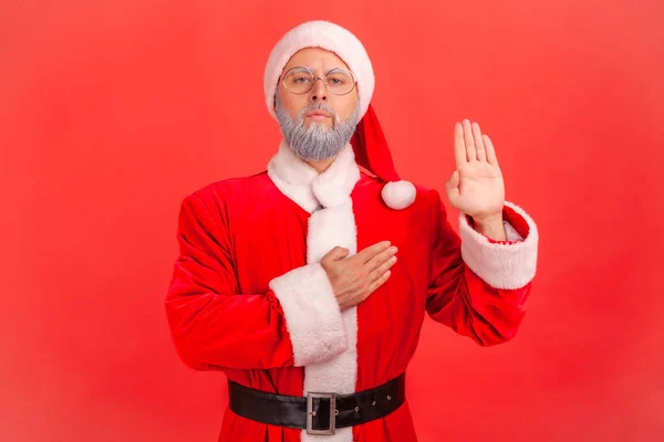 Serious elderly man with gray beard wearing santa claus costume standing raising hand and saying swear, making loyalty oath, pledging allegiance. Indoor studio shot isolated on red background.