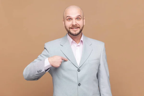 Portrait of bald bearded man pointing finger on himself and looking at camera with pride, extremely confident, wearing gray jacket. Indoor studio shot isolated on brown background.