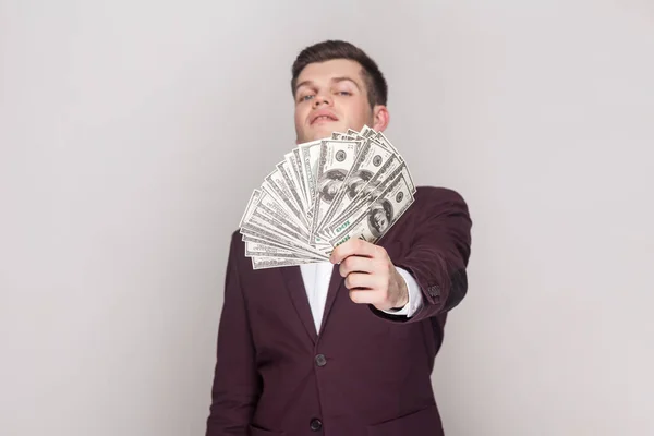 Portrait of arrogant wealthy man standing showing wad of money, looking at camera with proud expression, income, wearing violet suit and white shirt. Indoor studio shot isolated on grey background.