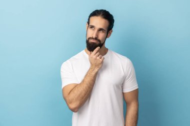 Beyaz tişörtlü, sakallı, düşünceli, yakışıklı bir adamın portresi. Çenesini tutuyor ve üzerinde kafa yoruyor. Çözüm konusunda kafası karışık. Mavi arka planda kapalı stüdyo çekimi.