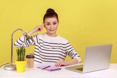 Ofis çalışanı aptal jestler yapıyor, parmaklarıyla kameraya bakıyor, yorgun bir ifadeyle iş yerinde dizüstü bilgisayarla oturuyor. Sarı arka planda kapalı stüdyo çekimleri.