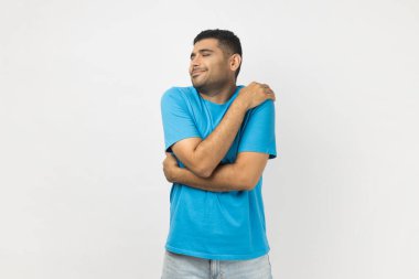 Mavi tişört giyen, mutlu, iyimser gülümseyen, kendine sarılan, tatmin olmuş ve mutluluğunu ifade eden bir adam portresi. Gri arka planda izole edilmiş kapalı stüdyo görüntüleri.