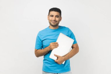 Mavi tişört giyen, elinde dizüstü bilgisayarla mutlu bir şekilde kameralara bakan iyimser ve iyimser gülümseyen bir adam. Gri arka planda izole edilmiş kapalı stüdyo görüntüleri.