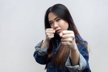 Kızgın, agresif, ciddi, esmer bir kadının portresi. Mavi kot ceketli, yumruklarını sıkmış, biriyle kavga ediyor. Gri arka planda izole edilmiş kapalı stüdyo görüntüleri.