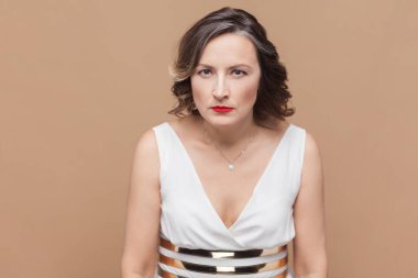 Öfkeli, orta yaşlı, dalgalı saçlı, beyaz elbiseli, nefretle kameraya bakan bir kadının portresi. Açık kahverengi arka planda kapalı stüdyo çekimleri.