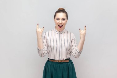 Heyecanlı, mutlu, çizgili tişört ve yeşil etek giyen, rock 'n roll jesti gösteren, festivalde eğlenen, çığlık atan bir kadının portresi. Gri arka planda izole edilmiş kapalı stüdyo görüntüleri.