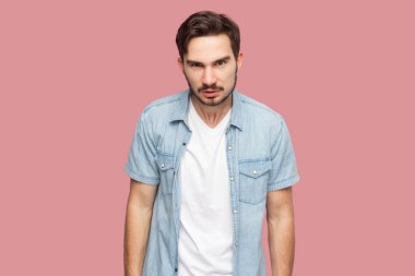 Mavi günlük gömlekli sakallı bir adamın portresi kaşlarını çatmış, suratında asık suratlı, birinin kötü davranışlarından memnun olmayan biri. Pembe arka planda kapalı stüdyo çekimleri.