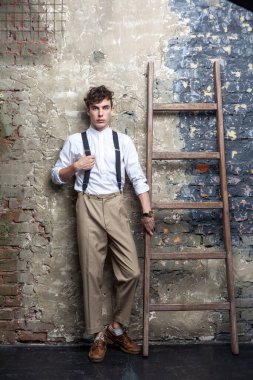 Beyaz gömlekli ve bej pantolonlu ciddi bir adamın eski ahşap merdivene dokunan, kameraya bakan, duvar arkasında poz veren uzun boylu portresi. Stüdyo içi çekim.