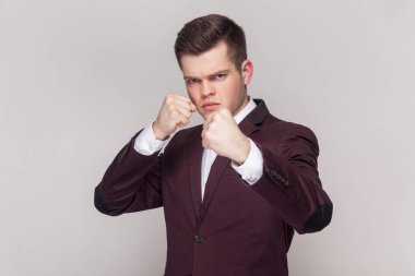 Yumrukları boksör yumruklu ve saldırıya ya da savunmaya hazır öfkeli bir yüzle kameraya saldırarak bakan mor takım elbiseli ve beyaz gömlekli bir adam. Kapalı alanda çekilen çekim gri arka planda izole edildi.