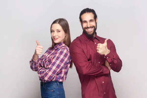Satisfied delighted woman and man standing together showing thumbs up, like gesture, positive feedback, looking at camera with happy expression. Indoor studio shot isolated on gray background.