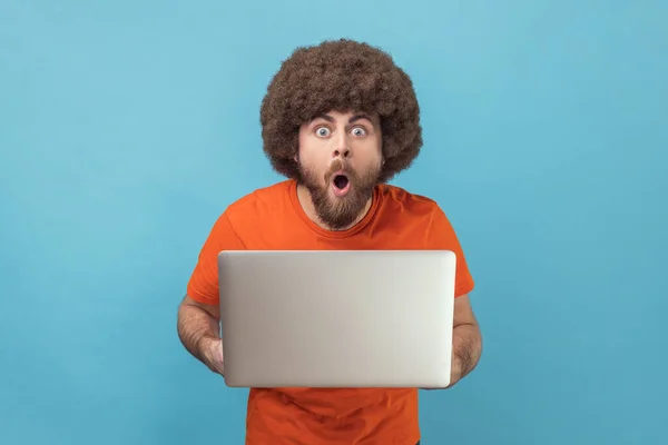 Portrait of astonished shocked man with Afro hairstyle wearing orange T-shirt standing holding laptop and looking at camera with open mouth and big eyes. Indoor studio shot isolated on blue background