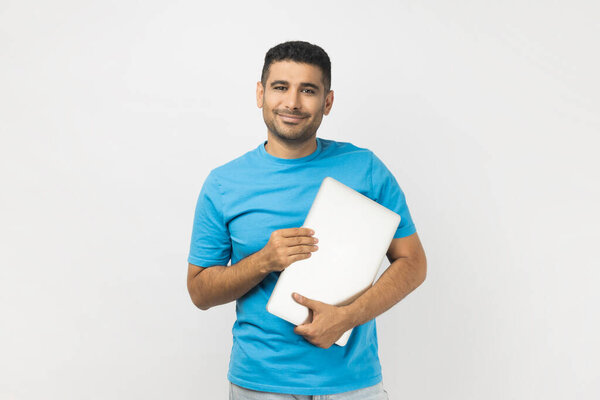 Улыбающийся оптимистичный небритый мужчина в голубой футболке, стоящий с закрытым ноутбуком, готовый к работе, с радостью смотрящий на камеры. Крытая студия, снятая на сером фоне.