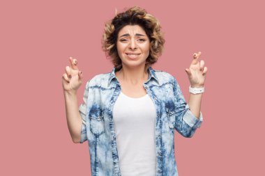 Kıvırcık saçlı, mavi gömlekli, parmakları çaprazlanmış ve kaşlarını çatmış güzel bir genç kadının portresi. Pembe arka planda kapalı stüdyo çekimleri.