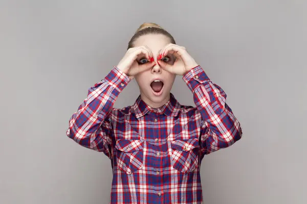 Portrait of amazed surprised excited woman with bun hairstyle making binocular gesture, looking at camera with open mouth, wearing checkered shirt. Indoor studio shot isolated on gray background.