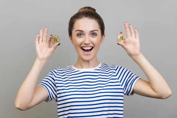 Çizgili tişört giyen, elinde bitcoin tutan, yeni dijital kripto para birimine dikkat eden heyecanlı bir kadının portresi. Gri arka planda izole edilmiş kapalı stüdyo görüntüleri.