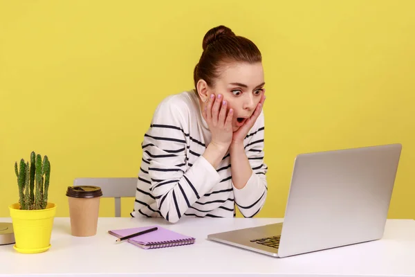 Surprised excited woman office manager in striped shirt looking at laptop with big eyes and open mouth, talking on video call. Indoor studio studio shot isolated on yellow background.