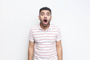 Şok olmuş, çizgili tişört giyen sakallı bir adamın portresi kameraya büyük ağızlı ve kocaman gözlü bakıyor ve hayret verici bir şey görüyor. Gri arka planda izole edilmiş kapalı stüdyo görüntüleri.