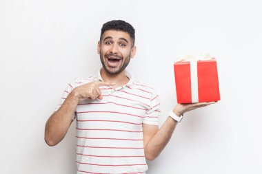 Şaşırmış, heyecanlı, sakallı, çizgili tişört giyen, elinde kırmızı ambalajlı hediye kutusu olan bir adamın portresi. Gri arka planda izole edilmiş kapalı stüdyo görüntüleri.